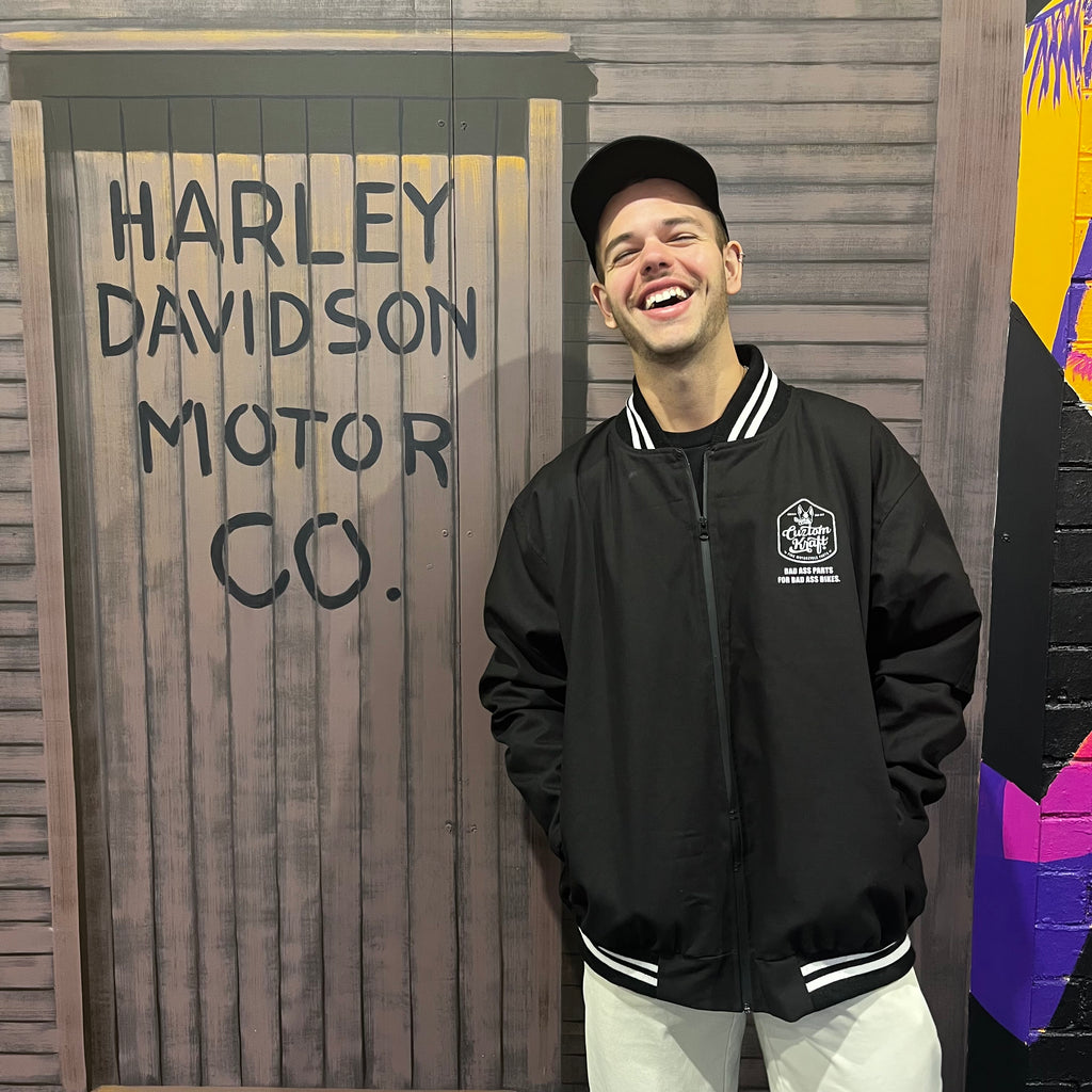 A man wearing a Cuztom Kraft 'Bad Ass' Jacket stands proudly in front of a mural that says Harley Davidson Motor Co.